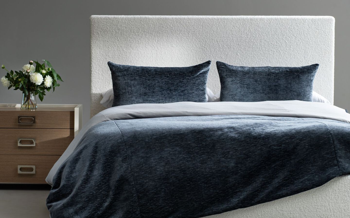 Bedroom with bed with grey upholstered bedframe and navy velvet comforter and a wooden nightstand on the side