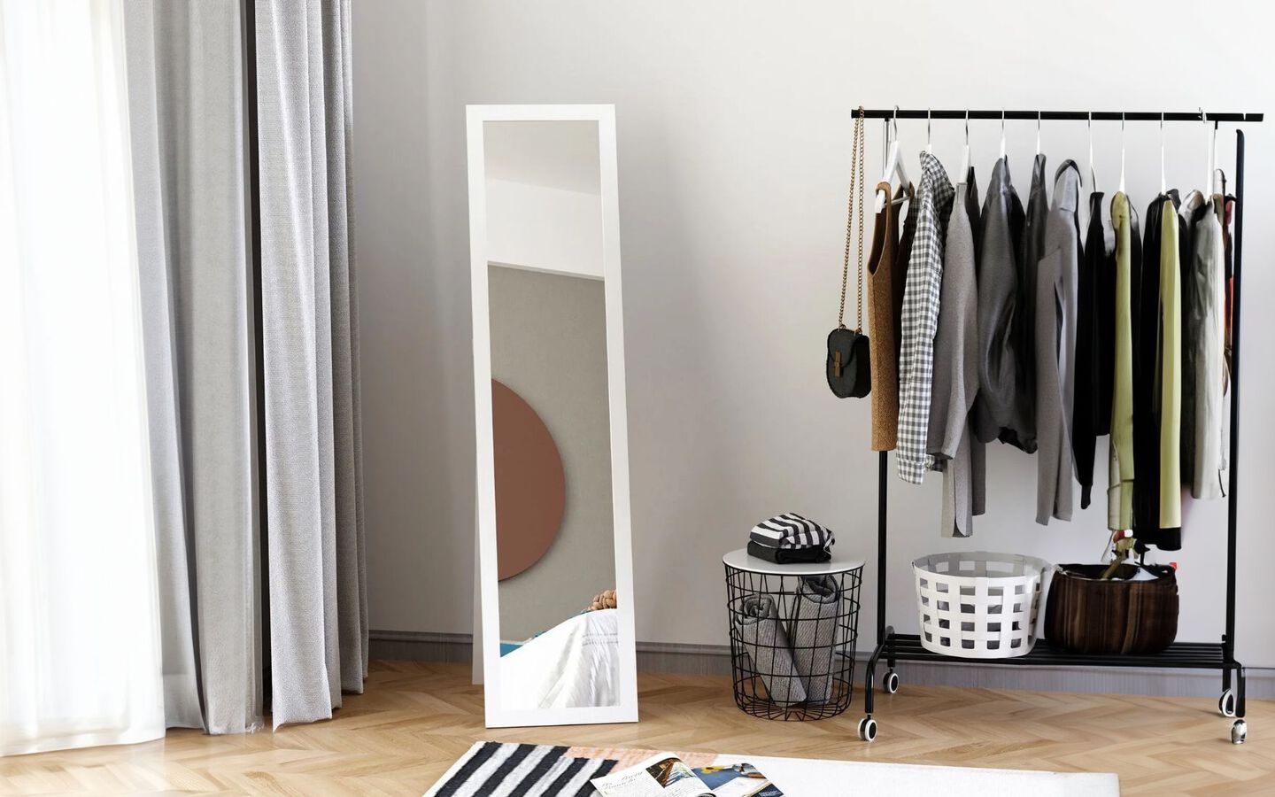 White standing mirror next to a rolling closet stand filled with clothes