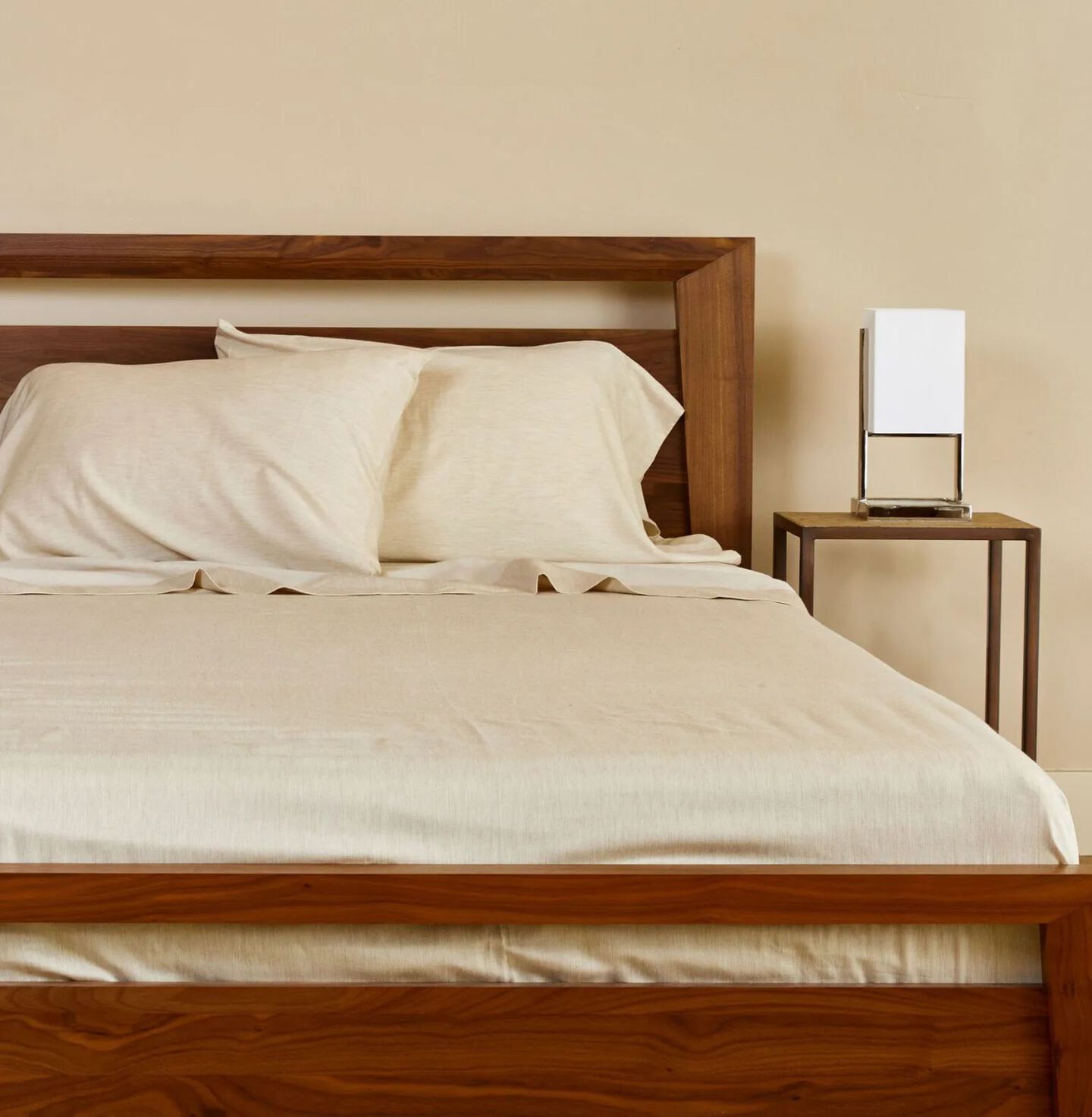 Bed with a dark wooden bedframe made with beige sheets next to a matching nightstand