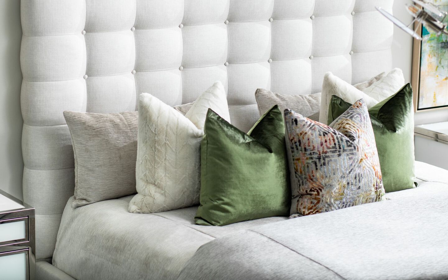 Closeup of bed with white upholstered bedframe, grey comforter, and green and grey pillows